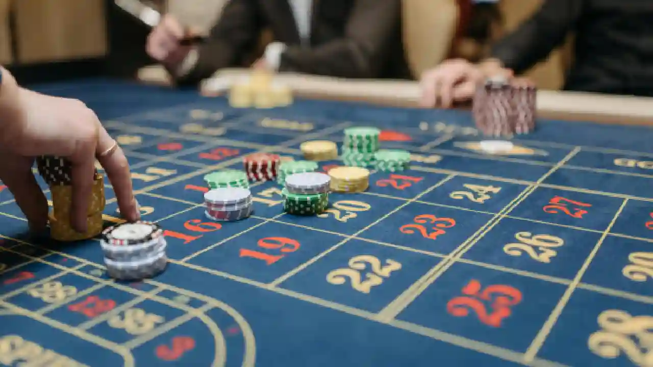 A close-up image of a roulette table in a casino. Chips of various colors are placed on the table, with a player's hand reaching to place a bet.