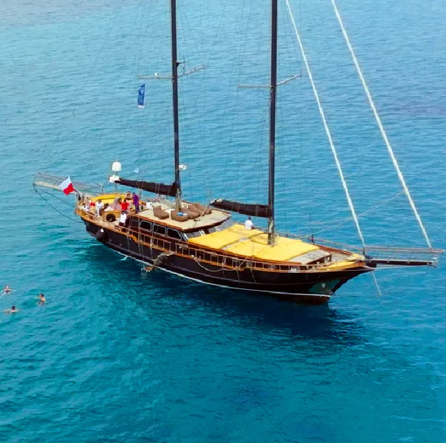 A large, traditional wooden gulet sailing yacht, painted in dark hues with wooden accents. The yacht is anchored in a crystal-clear turquoise bay, with people swimming and sunbathing in the water.