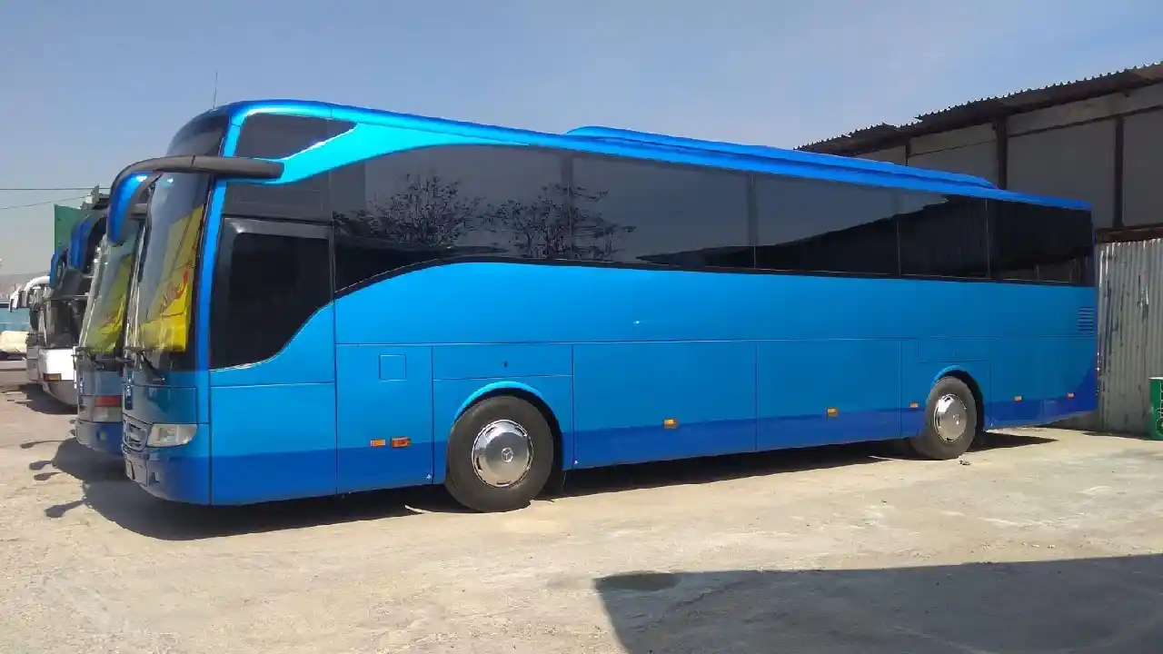 A large, blue Mercedes-Benz Tourismo coach bus parked in a parking lot near the Athens Bus Station. The bus has tinted windows and a sleek design, ready for long-distance travel.