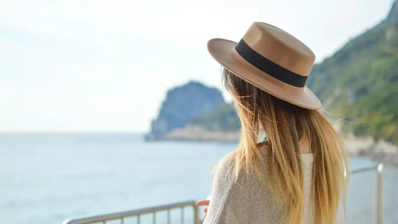 A woman wearing a hat stands at a beach in Greece, gazing out at the vast expanse of the Aegean Sea. The sun shines brightly, casting a warm glow on the water and the surrounding landscape.