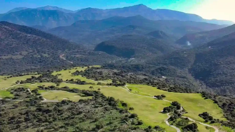 A breathtaking aerial view of a golf course nestled amidst rolling hills and mountains. The lush green fairways wind through the landscape, offering stunning views of the surrounding scenery.