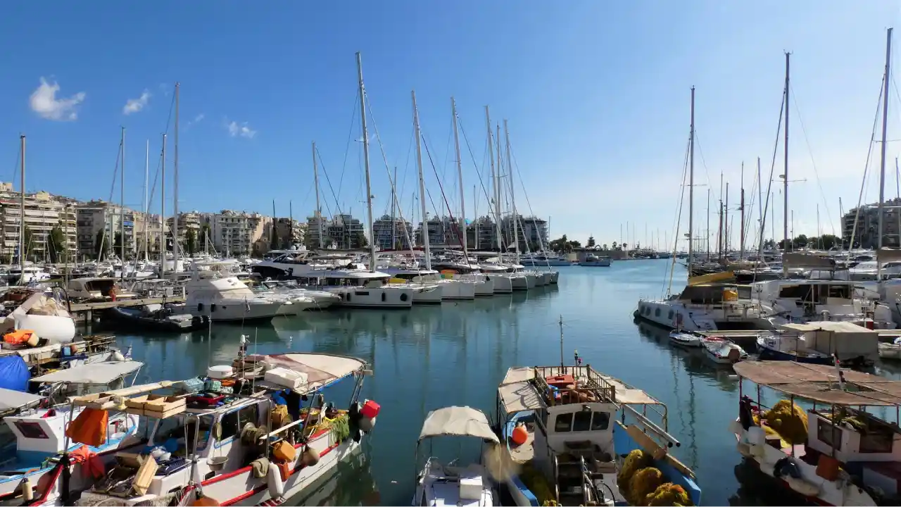 A panoramic view of Marina Zeas in Piraeus, Greece. Numerous yachts and sailboats are docked in the clear blue waters, surrounded by modern buildings and palm trees.