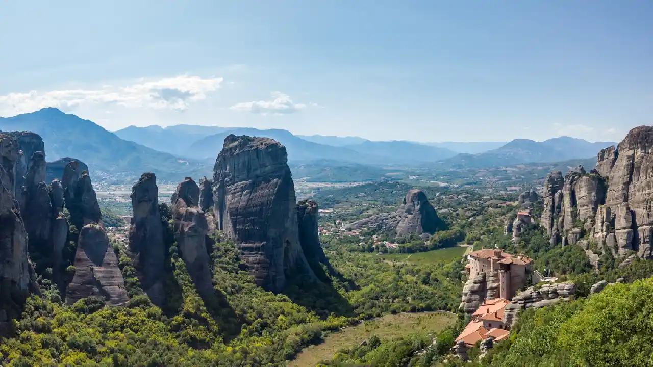 A breathtaking panoramic view of Meteora, Greece. Towering sandstone rock formations rise dramatically from a lush green valley, with ancient monasteries perched precariously on their peaks. The sky is a brilliant blue with fluffy white clouds.