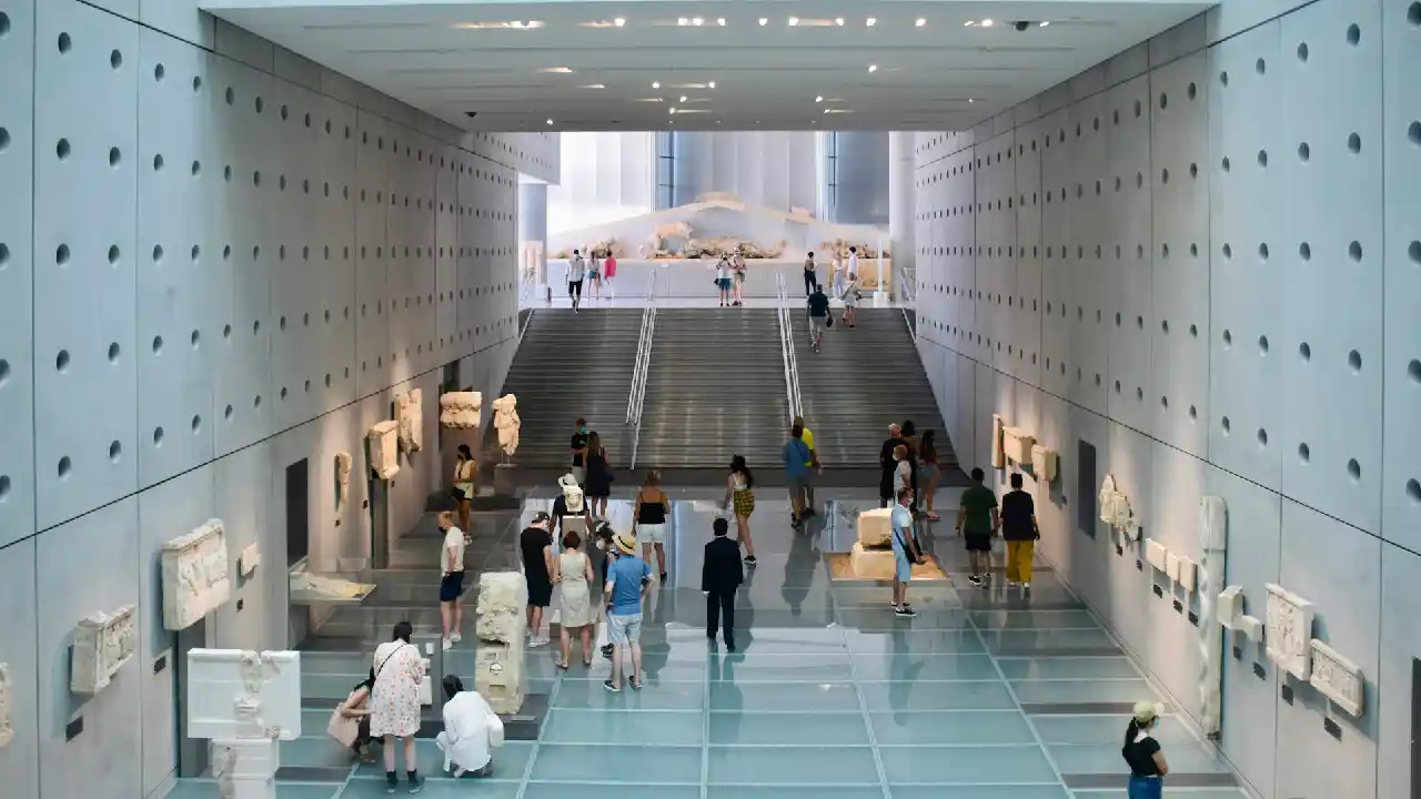 A photo of the Acropolis Museum in Athens, Greece. The museum building is a modern structure with a glass facade, blending seamlessly with the surrounding landscape.