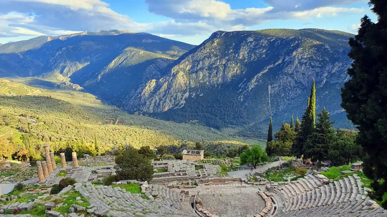 A panoramic view of the ancient Greek theater at Delphi. The theater is built into the hillside, with rows of stone seats overlooking a breathtaking valley and mountains in the distance.