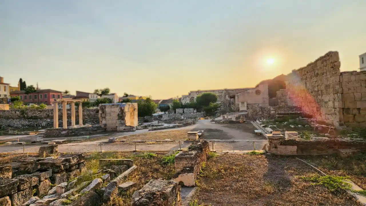 A panoramic view of the Roman Agora in Athens at sunset. The ruins of ancient temples and structures stand amidst a growing city, casting long shadows as the sun dips below the horizon.