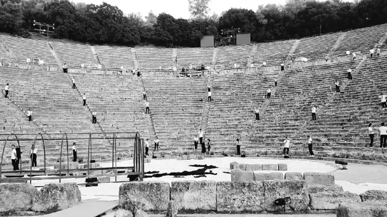 A black and white photograph of the ancient theater at Epidaurus, Greece. The stone steps of the amphitheater stretch upwards, creating a dramatic and awe-inspiring scene.