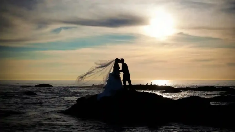 A silhouette of a bride and groom kissing on a rocky outcrop by the sea at sunset. The bride's veil flows dramatically in the wind, creating a romantic and ethereal scene.