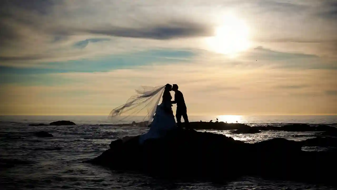 A silhouette of a bride and groom kissing on a rocky outcrop by the sea at sunset. The bride's veil flows dramatically in the wind, creating a romantic and ethereal scene.