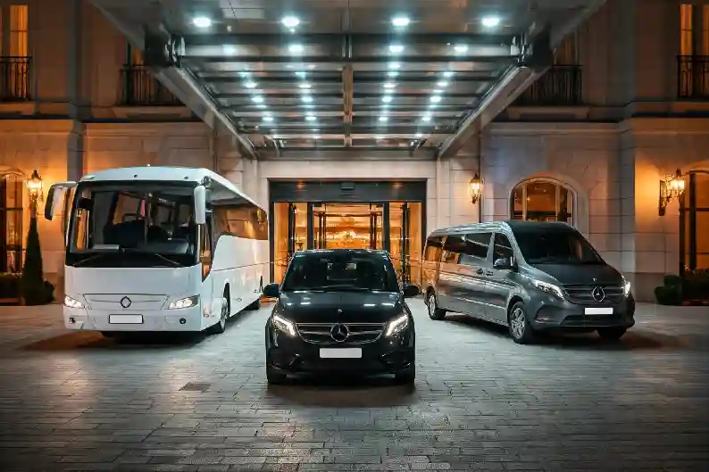 A luxury travel fleet parked in front of a grand hotel entrance. The fleet includes a sleek Mercedes-Benz car, a spacious minivan, and a comfortable coach bus, offering a range of transportation options for every travel need.