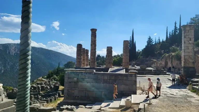 A photo of the Temple of Apollo at Delphi, Greece. The iconic temple ruins stand majestically against a backdrop of mountains and a clear blue sky. Visitors can be seen exploring the ancient site.