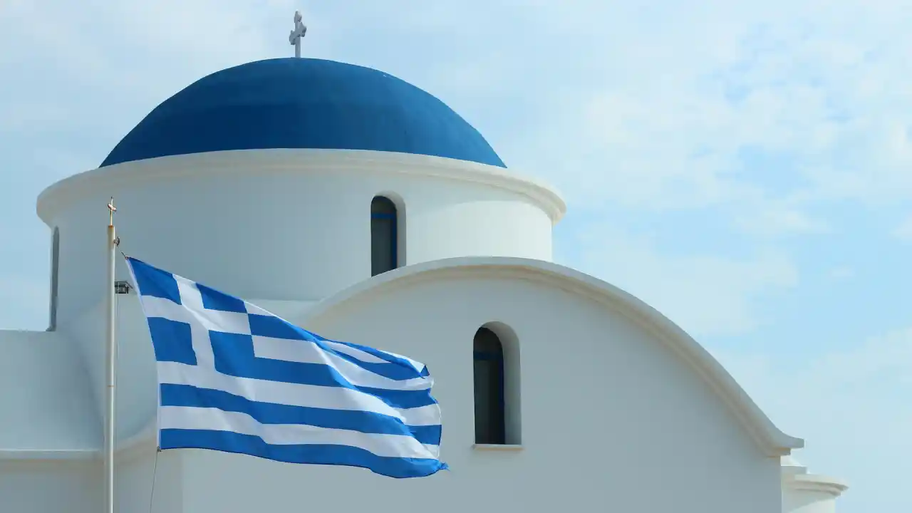 Welcome to Greece in Greek. A Greek Orthodox church with a blue dome and whitewashed walls stands against a clear blue sky. A Greek flag with its iconic blue and white stripes flutters proudly in the breeze.