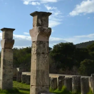 A row of ancient stone columns in Olympia with simple, square capitals stand against a backdrop of green trees and a blue sky with white clouds. The columns are weathered and show signs of age.