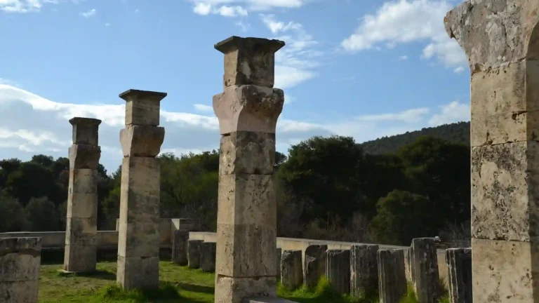 A row of ancient stone columns in Olympia with simple, square capitals stand against a backdrop of green trees and a blue sky with white clouds. The columns are weathered and show signs of age.