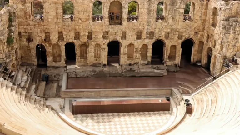 An overhead view of the Odeon of Herodes Atticus in Athens, Greece. The ancient Roman-era stone theatre features perfectly preserved semi-circular seating tiers leading down to the orchestra area, with the multi-story stone facade of the stage building visible in the background.
