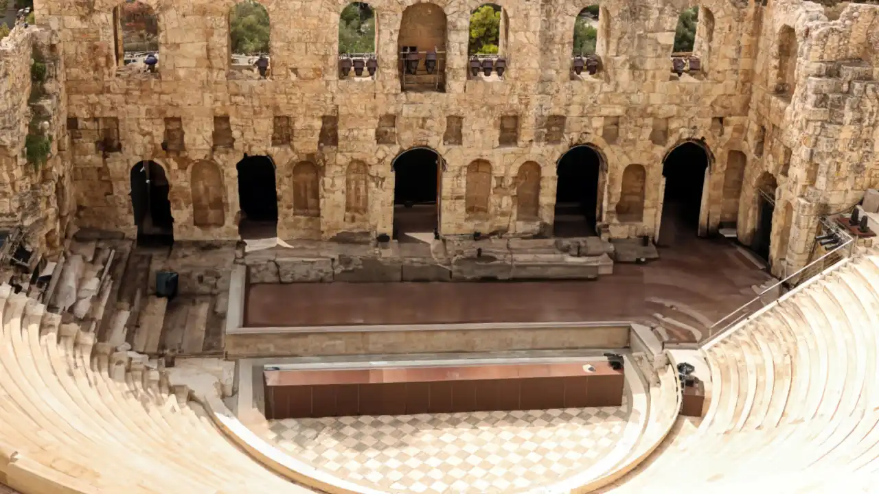 An overhead view of the Odeon of Herodes Atticus in Athens, Greece. The ancient Roman-era stone theatre features perfectly preserved semi-circular seating tiers leading down to the orchestra area, with the multi-story stone facade of the stage building visible in the background.