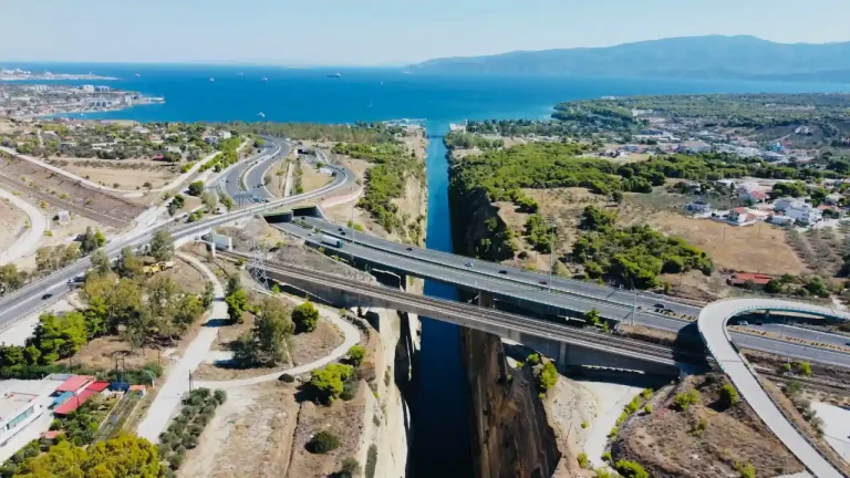 A high-angle aerial view of the deep, narrow Corinth Canal in Greece, featuring vibrant blue water. Multiple bridges, including a multi-lane highway and a railway line, span across the sheer limestone cliffs, with the surrounding coastal landscape and the sea visible in the distance.