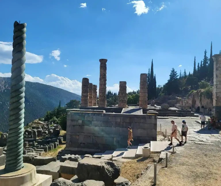 The ruins of the Temple of Apollo at Delphi, Greece, feature several standing Doric columns on a raised platform. The site is situated on a steep mountainside with a lush green valley in the background under a bright blue sky with scattered white clouds. Tourists are visible exploring the ancient pathways.
