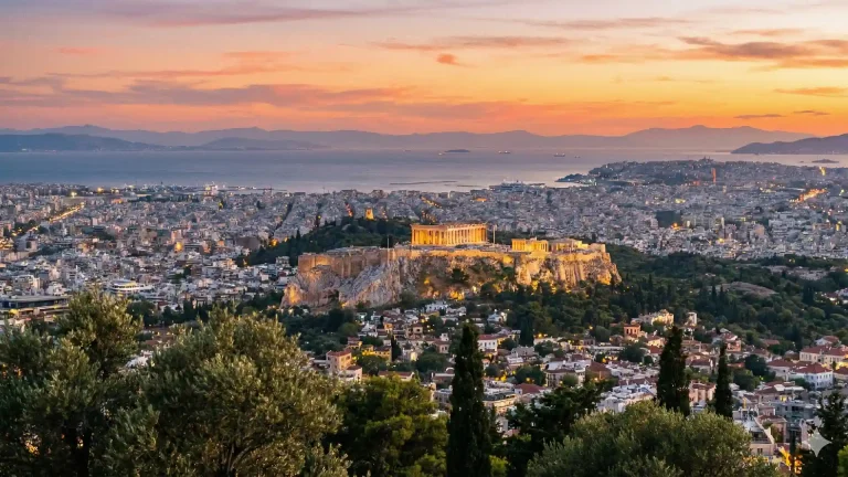 A panoramic landscape view of the Acropolis of Athens at sunset. The ancient Parthenon stands illuminated atop the rocky citadel, contrasting with the sprawling modern city grid below. The sky is painted in warm hues of orange and pink, with distant mountains framing the horizon.