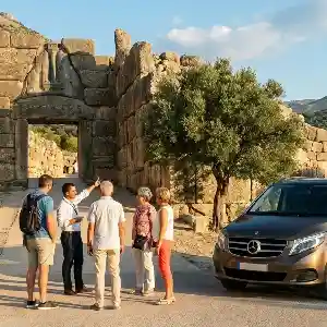 A professional tour guide explaining the history of the ancient Lion Gate at Mycenae to a small private group, with a luxury Mercedes tour van parked nearby under a clear blue sky.
