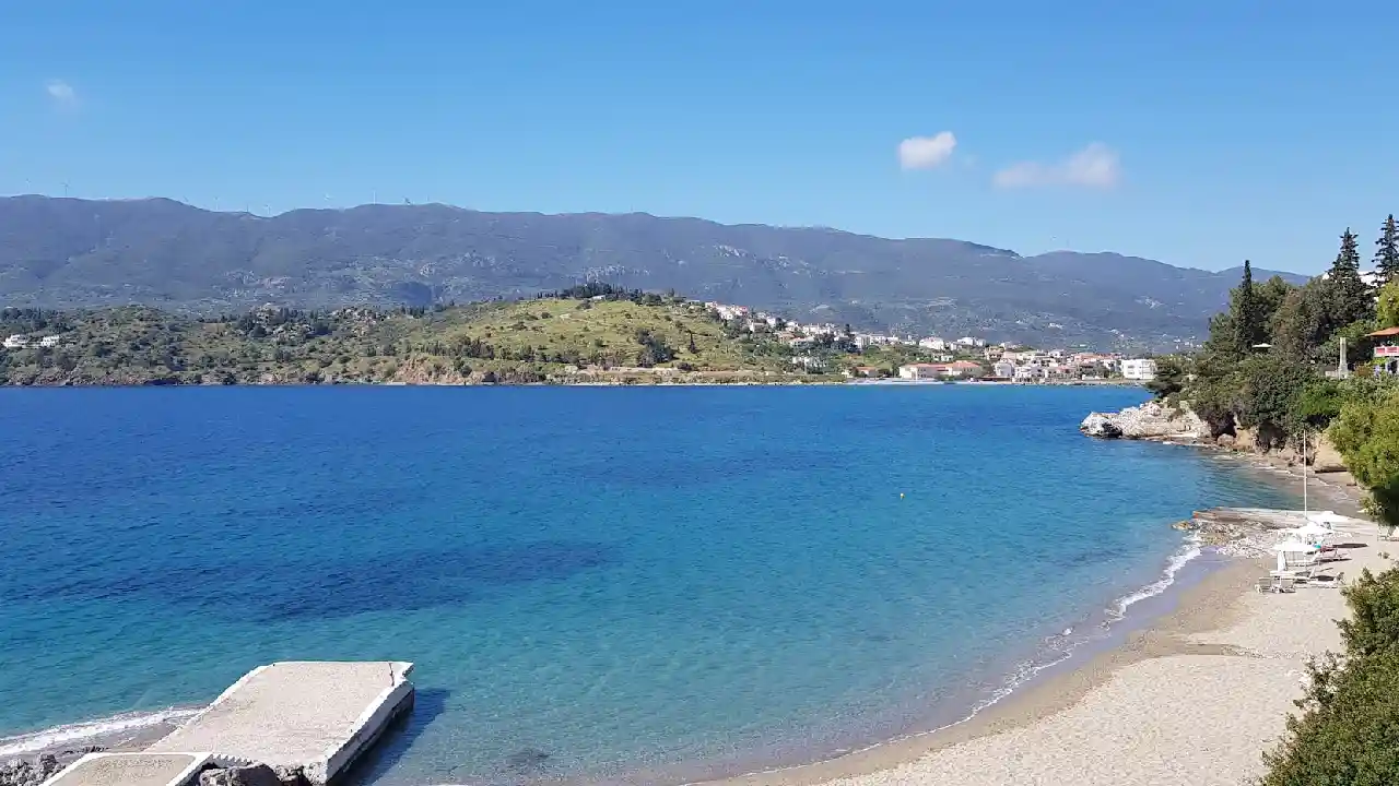 A scenic view of a tranquil sandy beach on Poros Island, Greece, featuring crystal clear turquoise water, a small stone pier, and lush green hills in the background.